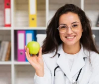 Médica sorridente com óculos de grau segurando uma maçã verde em um ambiente com prateleiras de livros ao fundo, promovendo saúde e bem-estar.