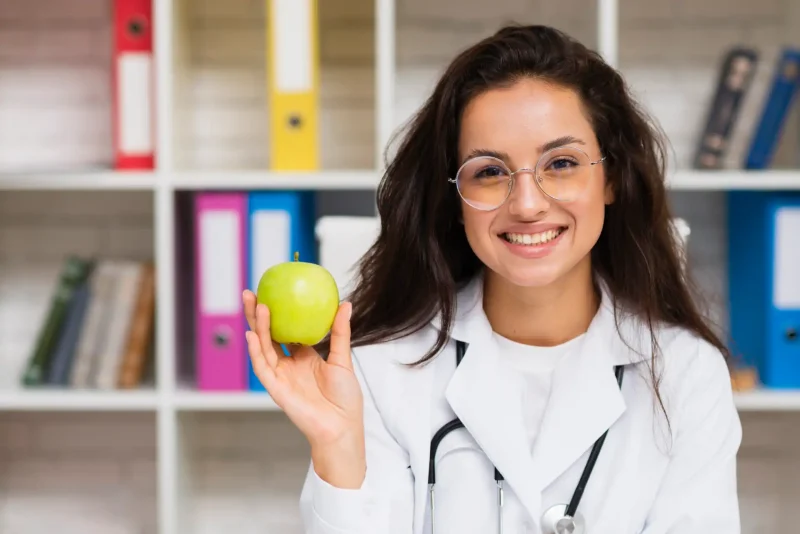 Médica sorridente com óculos de grau segurando uma maçã verde em um ambiente com prateleiras de livros ao fundo, promovendo saúde e bem-estar.