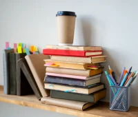 Imagem de uma mesa de estudos com várias pilhas de livros, uma caneca de papel para café ou chá, e uma gaveta com canetas coloridas, ideal para estudo ou trabalho.