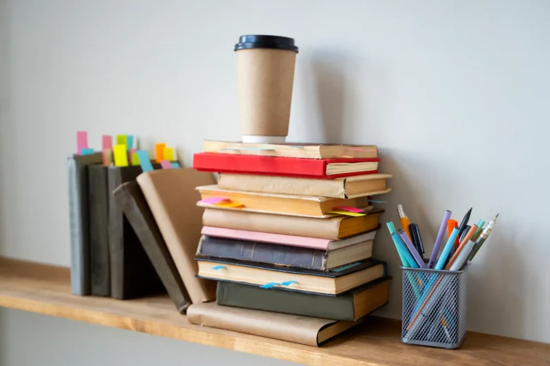 Imagem de uma mesa de estudos com várias pilhas de livros, uma caneca de papel para café ou chá, e uma gaveta com canetas coloridas, ideal para estudo ou trabalho.