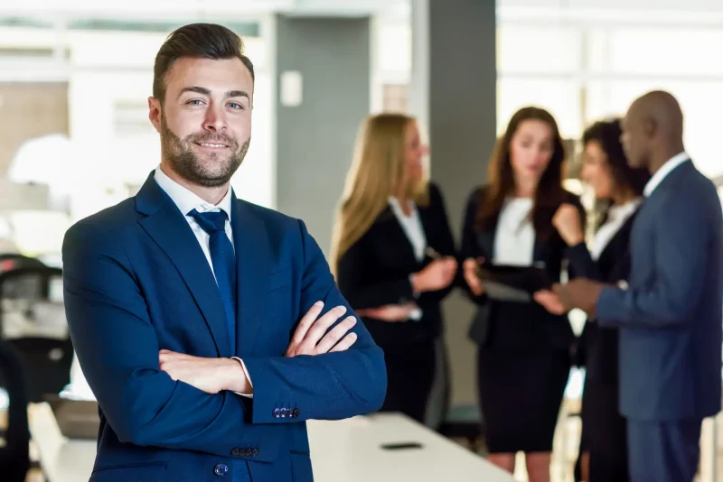 Homem de negócios sorridente com barba, usando terno azul escuro, em ambiente corporativo com colegas ao fundo. Imagem para otimização de SEO.