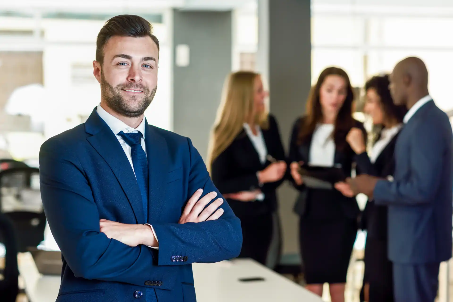 Homem de negócios sorridente com barba, usando terno azul escuro, em ambiente corporativo com colegas ao fundo. Imagem para otimização de SEO.
