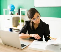 Mulher estudando e preparando um plano de aula em sala de aula com caderno, laptop e livros na mesa.