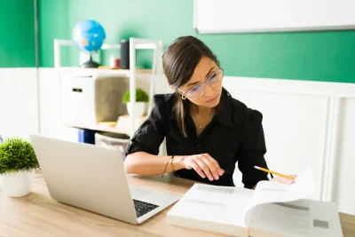 Mulher estudando e preparando um plano de aula em sala de aula com caderno, laptop e livros na mesa.