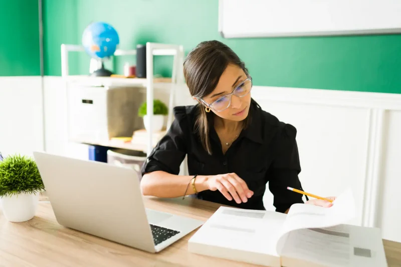 Mulher estudando e preparando um plano de aula em sala de aula com caderno, laptop e livros na mesa.