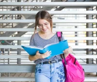 Estudante universitária com mochila rosa lendo livro ao ar livre, simbolizando sucesso na universidade e alta nota no ENEM.