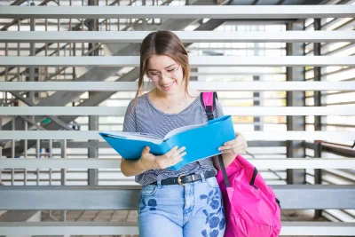 Estudante universitária com mochila rosa lendo livro ao ar livre, simbolizando sucesso na universidade e alta nota no ENEM.