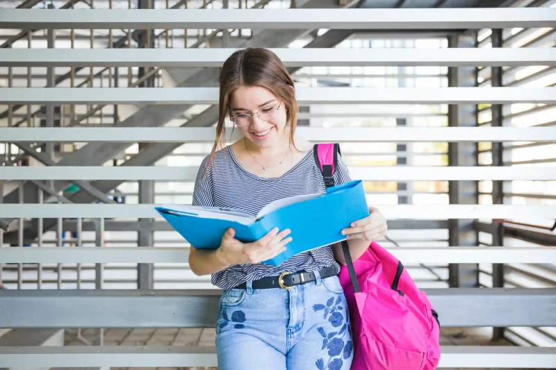 Estudante universitária com mochila rosa lendo livro ao ar livre, simbolizando sucesso na universidade e alta nota no ENEM.