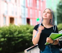 Jovem estudante universitária pensando em nota do ENEM enquanto segura caderno na mão ao ar livre, em frente a um prédio universitário.