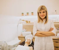 Esteticista sorridente com braços cruzados em clínica de estética, atendendo uma cliente relaxada na cadeira de tratamento facial, ambiente clean e bem iluminado