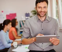 Profissional de marketing digital em ambiente de escritório com colegas ao fundo, usando tablet e sorrindo.