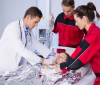 Equipe médica e de enfermagem em hospital, com profissional usando estetoscópio examinando paciente deitado e equipe ao lado ajudando no procedimento, em ambiente hospitalar.
