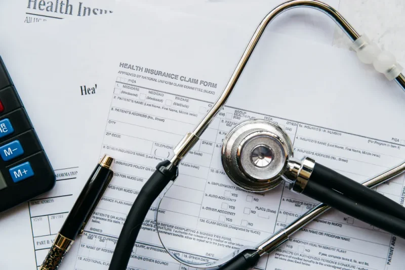 Formulário de seguro saúde com carimbo “Health Insurance Claim Form”, canetas e estetoscópio sobre uma mesa, sugerindo atendimento médico, sinistro e documentação para reembolso.