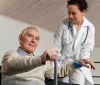 Médica enfermeira auxilia um idoso sentado em casa, oferecendo apoio para caminhar com andador e orientando o movimento durante atendimento de saúde, com luvas azuis.