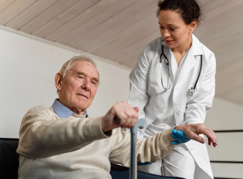 Médica enfermeira auxilia um idoso sentado em casa, oferecendo apoio para caminhar com andador e orientando o movimento durante atendimento de saúde, com luvas azuis.