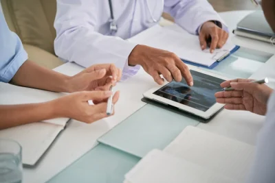 Equipe em reunião com profissionais de saúde revisando informações em tablets e segurando canetas, sugerindo planejamento e consulta médica em ambiente clínico.