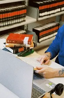 Uma pessoa em uma mesa na biblioteca com livros e um notebook pesquisando.