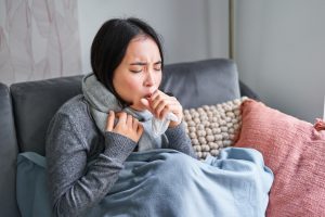 Asian woman coughing, feeling sick, sitting under blanket with scard in cold apartment, saving on heating this winter, catching flu.