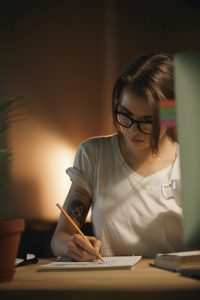 Picture of serious young woman designer sitting indoors at night writing notes using computer. Looking aside.